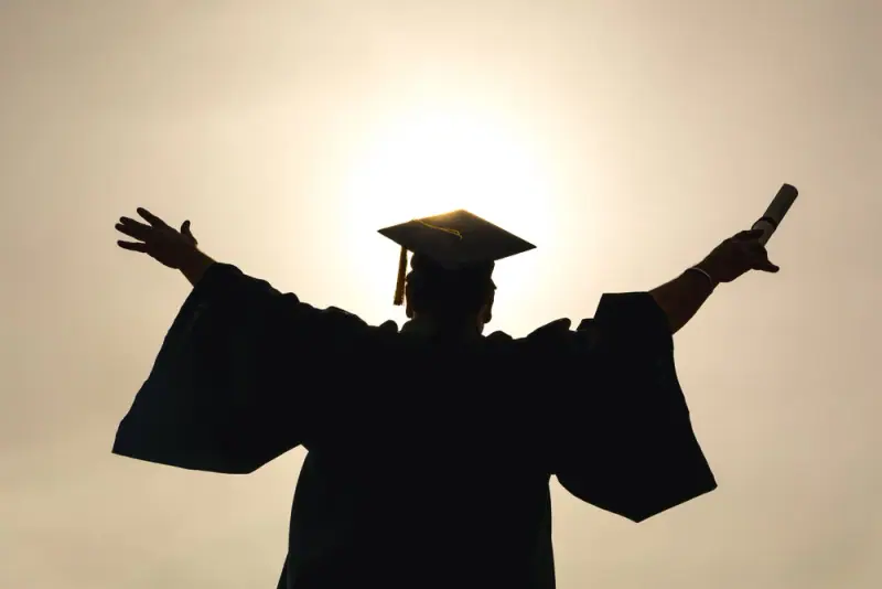 Silhouette of a graduate raising their arms and diploma against a golden sky